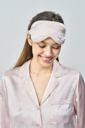 Smiling model with lowered gaze wears pink Alice Daisy Eye Mask embroidered with daisies, evoking gentle comfort and a cozy vibe.