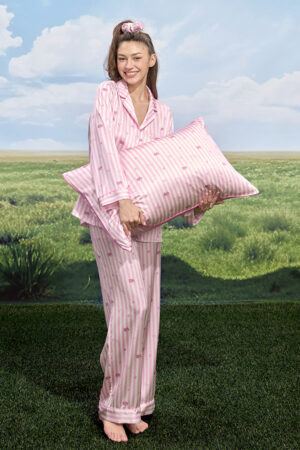 Smiling model holding large pillow from MANITO DISNEY Cheshire Collection Sleeping Gift Box in matching pink striped silk pajamas.