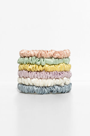 Stack of six skinny silk scrunchies in pastel shades of peach, yellow, green, purple, cream, and blue displayed on a bright white background.