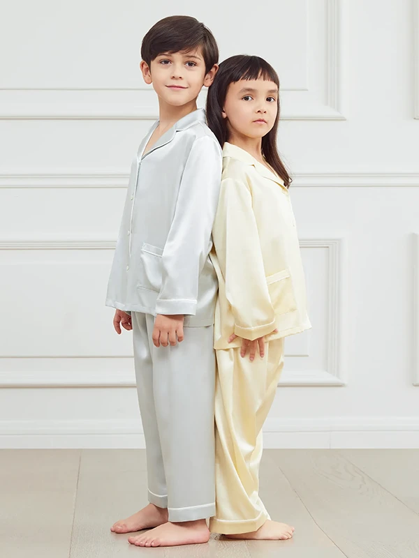 Two kids in MANITO silk pajama sets standing back-to-back, dressed in soft silver and cream tones.